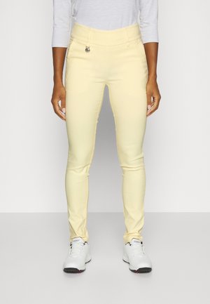 Person trägt schlanke hellgelbe Hose, weißes langärmeliges Oberteil und weiße Sneakers und steht vor einem schlichten Hintergrund.