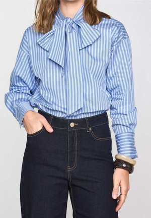 Frau trägt eine blau gestreifte Bluse mit großer Schleife und dunkle High-Waist-Jeans, Hand in der Tasche und klobige Armbänder am Handgelenk.