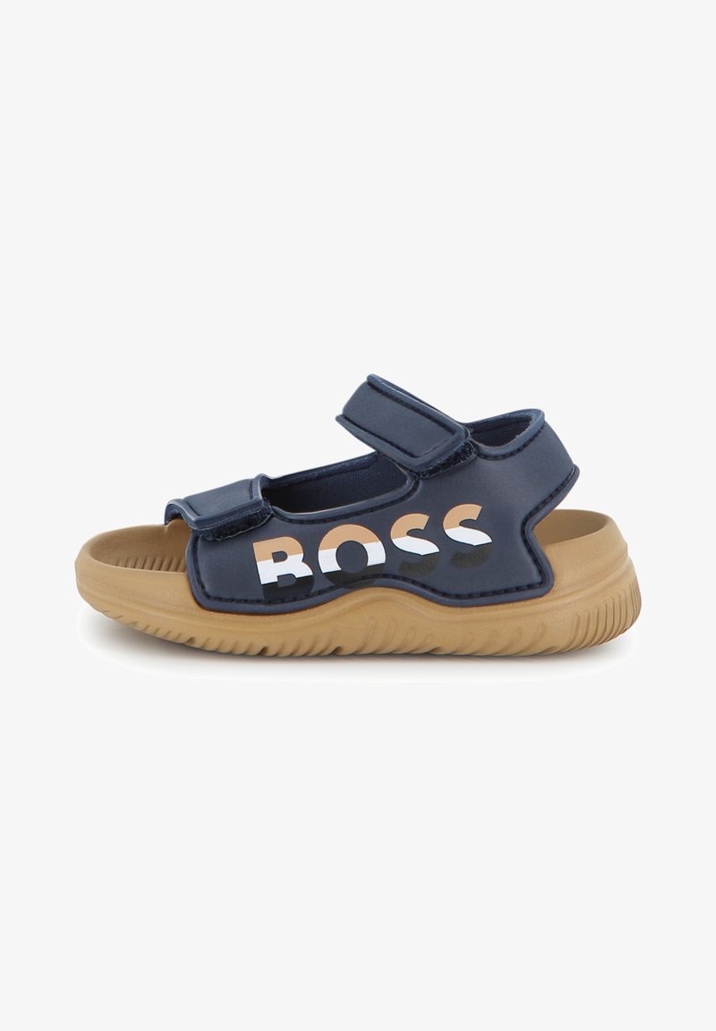 Marineblauwe sandalen met klittenband, een tankleurige rubberen zool, met een opvallend "BOSS" logo in wit en beige aan de zijkant.