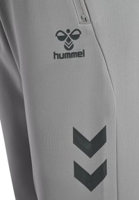 Graue Sporthose mit schwarzem "hummel"-Logo und Chevron-Muster, mit glatter Textur und Reißverschlusstasche.