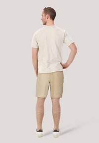 Ljust beige bomullströja med korta ärmar, i kombination med ljusbruna shorts och vita sneakers med gröna detaljer. Sedd bakifrån.