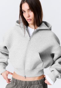 Ung kvinne i cropped lys grå hettejakke med glidelås over en hvit skjorte, justerer mørkegrå bukser med elastisk midje mot en ensfarget hvit bakgrunn.