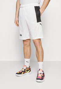 Pantalones cortos deportivos blancos con un panel lateral de malla negra y el logo de PUMA. Zapatillas deportivas negras y multicolores con un patrón vibrante y calcetines blancos.