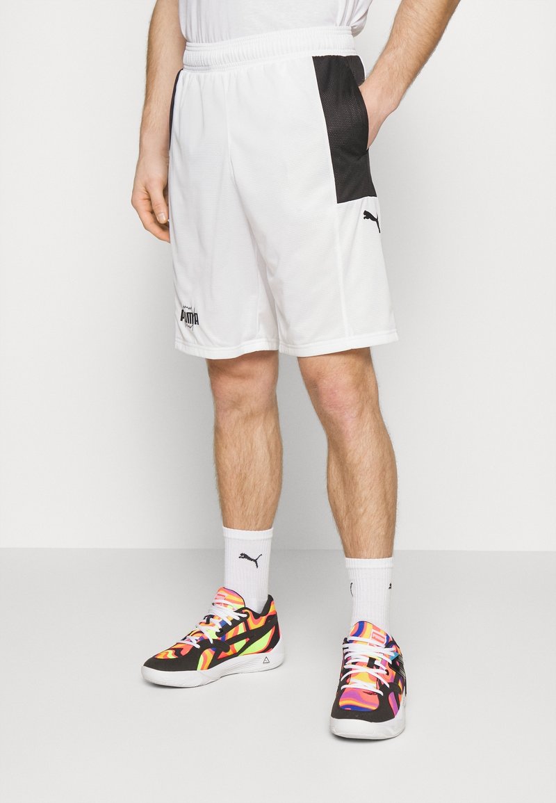Pantalones cortos deportivos blancos con un panel lateral de malla negra y el logo de PUMA. Zapatillas deportivas negras y multicolores con un patrón vibrante y calcetines blancos.