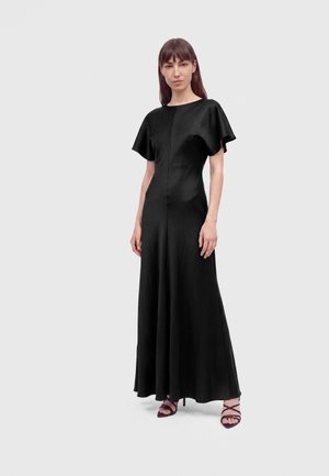 Femme debout portant une longue robe en satin noire avec des manches courtes à volants et des sandales noires à talons hauts à brides, sur un fond uni.