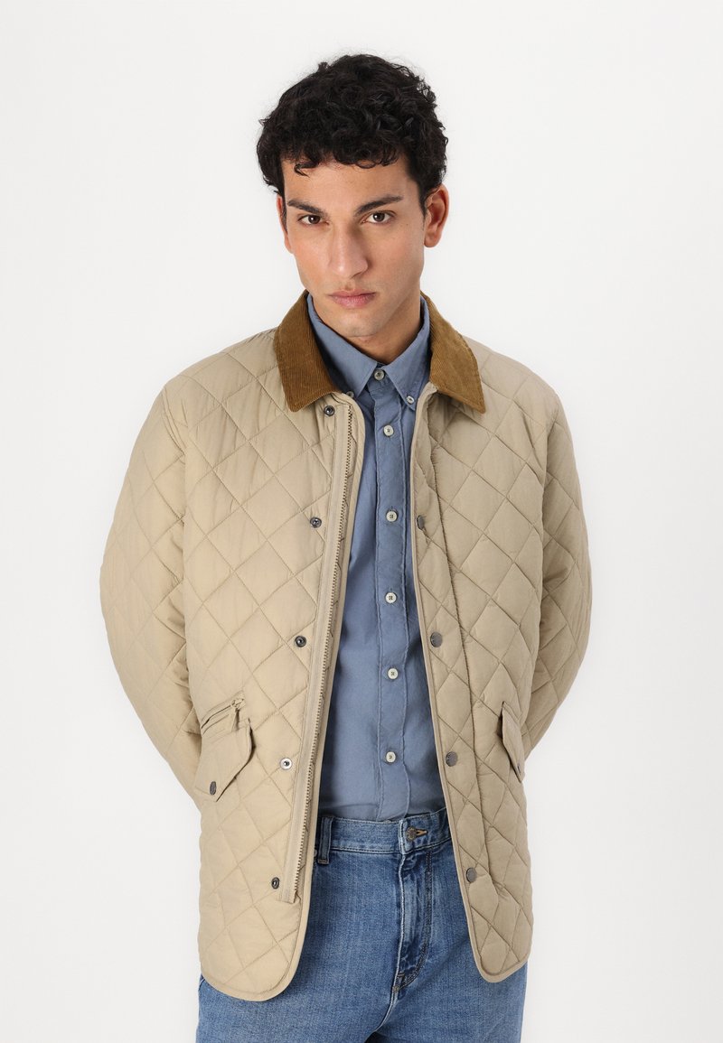 Beige gesteppte Jacke mit einem braunen Cordkragen, Druckknöpfen und Seitentaschen, getragen über einem hellblauen Hemd und Jeans.