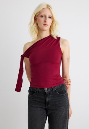 Kvinde med blondt hår iført ærmeløs burgunderfarvet top med asymmetrisk skulderbind og sorte jeans, stående mod en ensfarvet baggrund.