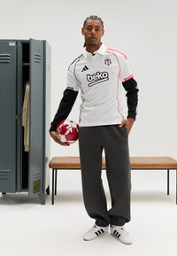 Camisola de futebol branca e preta com detalhes em vermelho, calças de treino cinzentas e sapatilhas desportivas brancas, segurando uma bola de futebol com padrão vermelho e branco.