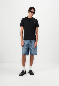 Schwarzes Baumwoll-T-Shirt mit kurzen Ärmeln und Logo, blaue gemusterte Jeansshorts, weiße Socken und schwarze Slipper. Das Modell steht vor einem weißen Hintergrund.
