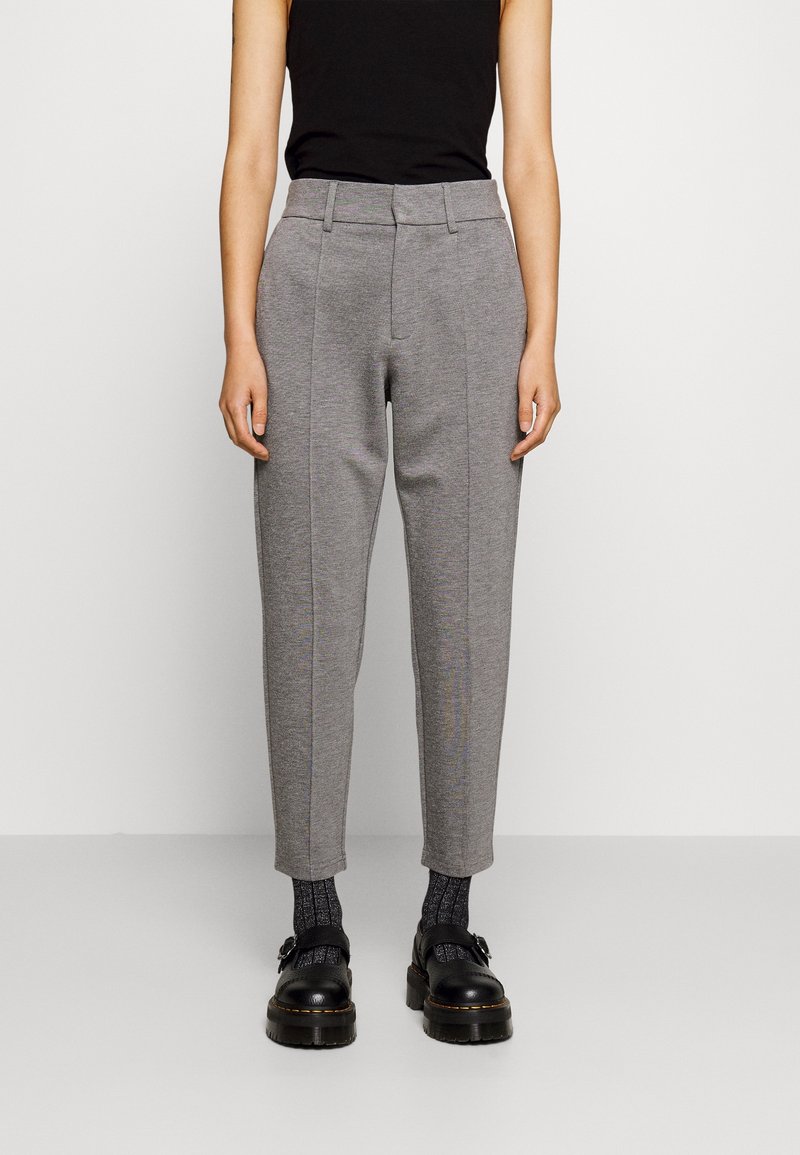 Personne portant un pantalon gris sur mesure longueur cheville, des chaussettes noires à motifs, des chaussures noires épaisses et un haut noir sans manches, debout devant un fond uni.
