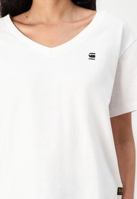 Bijela pamučna majica s V-izrezom, kratkim rukavima i malim crnim logotipom na lijevoj strani prsiju. Ima teksturiranu tkaninu i božni natpis.