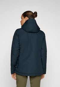 Veste isolante bleu foncé avec capuche, présentant une texture lisse, des coutures d'épaule et un petit logo près de la manche gauche.
