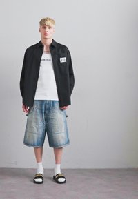 En blond mand står op ad en simpel væg iført en sort åben skjorte, en hvid tanktop, løse denimshorts, hvide sokker og sorte slides med gul tekst.