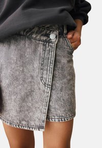 Persona che indossa una mini gonna in denim acid washed con una mano in tasca, abbinata a una maglietta scura a maniche lunghe, su uno sfondo neutro.