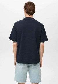 Tricou din bumbac de culoare bleu marin, cu mâneci scurte, croială lejeră, guler rotund și textură netedă. Are spatele simplu. Este purtat cu pantaloni scurți de culoare bleu deschis.