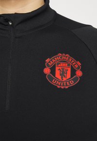 Camisa negra con cremallera que presenta un prominente escudo rojo del Manchester United en el pecho, con detalles y tela texturizada.