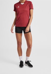 Camiseta atlética color carmesí con cuello en V y mangas cortas, combinada con pantalones cortos negros. Presenta el logotipo de la marca en el pecho y un diseño deportivo.