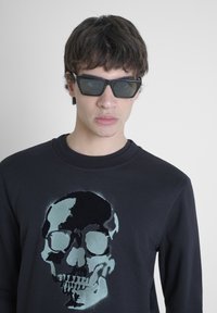 Un sweat-shirt noir avec un grand graphisme de crâne abstrait dans des nuances de gris et de turquoise. Le mannequin porte des lunettes de soleil noires angulaires.