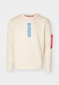 Puha anyagból készült bézs pulóver, kék téglalap alakú "ALPHA" logóval és piros cipzáras zsebbel a bal ujjon.