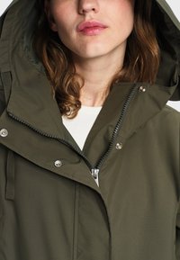 Femme portant une veste à capuche vert olive avec une fermeture éclair et des boutons-pression, une chemise blanche visible en dessous, et des cheveux bruns lâches.