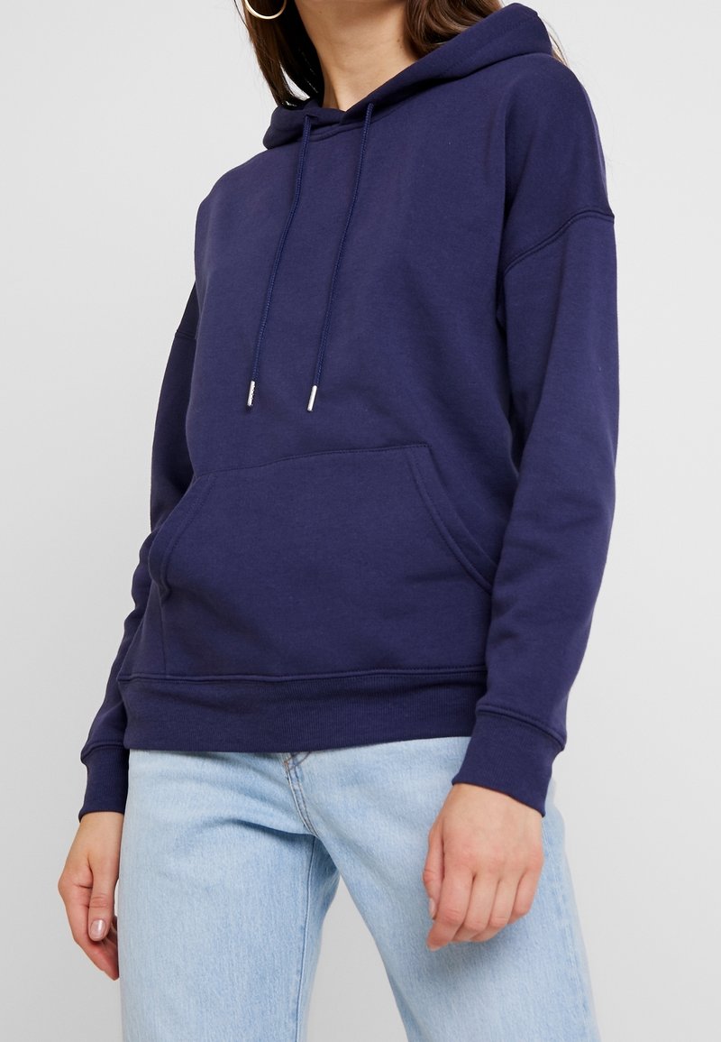 Femme portant un sweat à capuche bleu foncé avec une poche avant et un jean bleu clair, sur un fond clair uni.