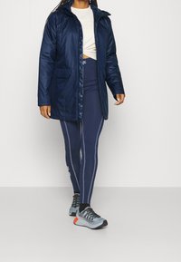 Marinblå pufferjacka med huva, med en framsida som har dragkedja och sidofickor. Matchad med marinblå leggings och flerfärgade träningstejpor.