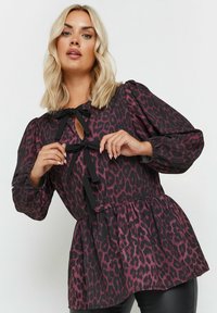 Pitkähihaisessa mustassa paidassa on violetti leopardi-printti, jossa on avainaukko-aukko kauluksessa ja mustat nauhat kaulalla.