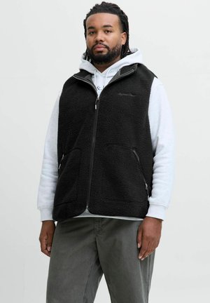 Mand med dreadlocks iført en sort lynlås fleecevest over en lysegrå hoodie og mørkegrå bukser, stående mod en ensfarvet baggrund.