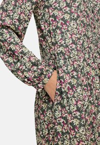 Bloemenjurk van donkergroene stof met witte en roze bloemenpatronen, lange mouwen met gerimpelde manchetten en zijzakken.