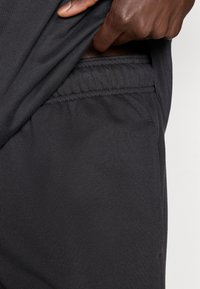 Μαύρα αθλητικά σορτς με ελαστική μέση, που διαθέτουν πλαϊνή τσέπη και λεία υφή υφάσματος, που είναι εν μέρει μισοσκεπασμένα  κάτω από ένα πουκάμισο.