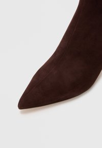 Botín de ante marrón con punta afilada, textura suave y diseño sin costuras, que presenta un tacón alto y sin hardware visible.