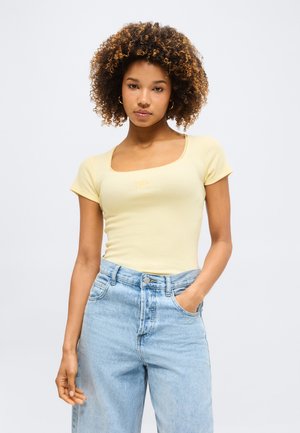 Νεαρή γυναίκα με σγουρά μαλλιά που φοράει ένα ανοιχτό κίτρινο εφαρμοστό τοπ με κοντά μανίκια και ανοιχτό μπλε ψηλόμεσο τζιν, με το ένα χέρι στην τσέπη.