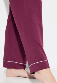 Pantaloni da pigiama bordeaux realizzati in tessuto morbido, caratterizzati da una vestibilità comoda, orli con piping bianco e lunghezza che scopre le caviglie.