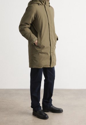 Mann trägt eine olivgrüne Winterjacke mit Kapuze, marineblaue Hose und schwarze Schuhe, steht mit den Händen in den Jackentaschen vor einem schlichten Hintergrund.