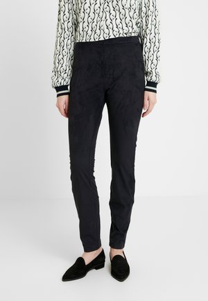 Pantalons noirs ajustés en matériau doux, ressemblant à du daim. Dotés d'une texture lisse et d'une taille mi-haute. Portés avec des chaussures noires sans lacets.