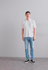 Witte polo met korte mouwen van een gestructureerde stof, gecombineerd met lichtblauwe jeans en zwarte sneakers met witte zolen. Effen achtergrond.