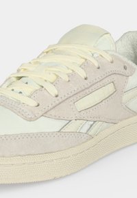 Близък план на кремаво и бежово маратонка Reebok с велурени наслагвания, кремави връзки и текстурирана подметка на светъл фон.