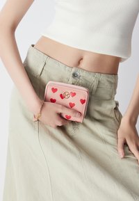 Roze portemonnee met rode hartpatronen, gouden hardware logo, afgebeeld vastgehouden tegen een lichtgroene rok, met een aansluitend wit topje.
