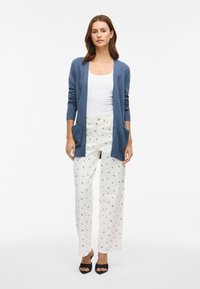 Μπλε πλεκτό ζακέτα με μακριά μανίκια και πλαϊνές τσέπες, συνδυασμένη με κρεμ παντελόνι με λουλουδάτο μοτίβο. Μαύρες ανοιχτές γόβες ολοκληρώνουν την εμφάνιση.