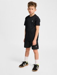 Svart t-shirt och shorts-set med en elegant design; t-shirten har en liten vit logotyp, texturerade sneakers, vita strumpor och kontrasterande skodetaljer.