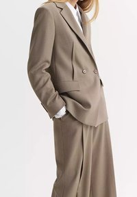 Femme portant un blazer beige croisé avec un pantalon large assorti et une chemise blanche, les mains dans les poches, sur un fond uni.