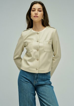 Mujer con cabello castaño suelto que lleva una chaqueta beige abotonada con bolsillos en el pecho y vaqueros azules de tiro alto, de pie frente a un fondo claro y liso.