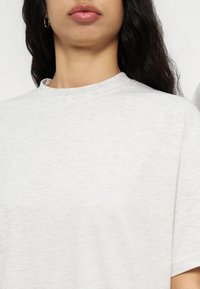 Γυναίκα με μακριά σκουρόχρωμα μαλλιά, που φοράει ασημένια σκουλαρίκια κρίκους και απλό ανοιχτό γκρι t-shirt με στρογγυλή λαιμόκοψη, εμφανιζόμενη από τους ώμους έως τα χείλη.