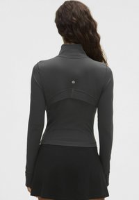 Mujer con cabello largo y ondulado que lleva una blusa ajustada de manga larga gris oscuro y una falda negra acampanada, vista desde atrás sobre un fondo liso.