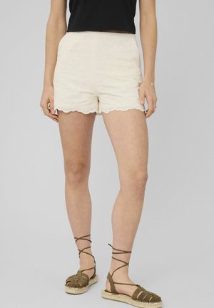 Femme portant un short en dentelle crème à ourlets festonnés, un haut noir, et des sandales espadrilles kaki à lacets à la cheville, debout devant un fond uni.