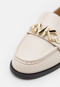 Mocassin en cuir crème avec semelle noire et détail du logo MK en métal doré sur la bride à l'avant.