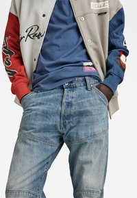 Herenoutfit met een grijs-rode jas met logo's, een blauwe t-shirt en lichtblauwe jeans met een vervaagde textuur en een patchlabel.