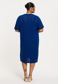 Blaues Strickkleid mit geometrischem Muster, kurzen Ärmeln und knielanger Länge. Texturiertes Material und offene Details sorgen für visuelles Interesse.