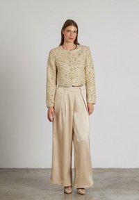 Beige Tweed-Jacke mit strukturiertem Finish, rundem Ausschnitt und Fronttaschen. Kombiniert mit beigefarbenen Satin-Hose im weit geschnittenen Stil und passenden Schuhen.