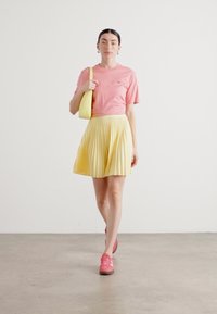 T-shirt di cotone rosa, gonna plissettata gialla e sneakers rosa. La modella porta una borsa a tracolla gialla, in piedi su un pavimento grigio texturizzato.